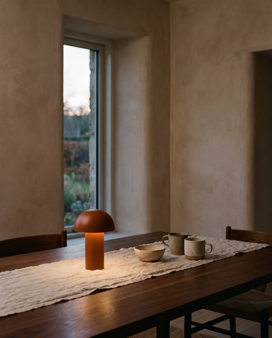 A rust-coloured cordless mushroom lamp glowing on a walnut dining table indoors at dusk