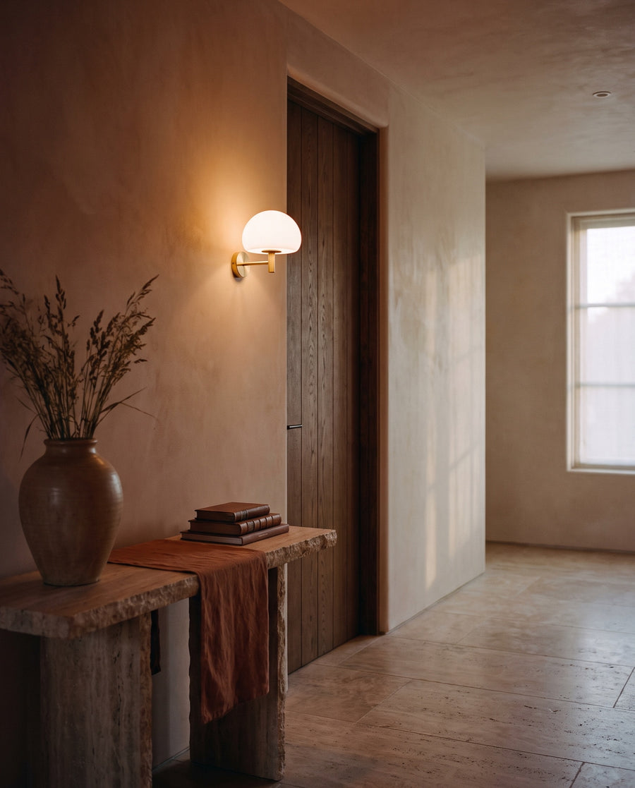 An opal glass wall sconce glowing beside an oak door in a plaster hallway