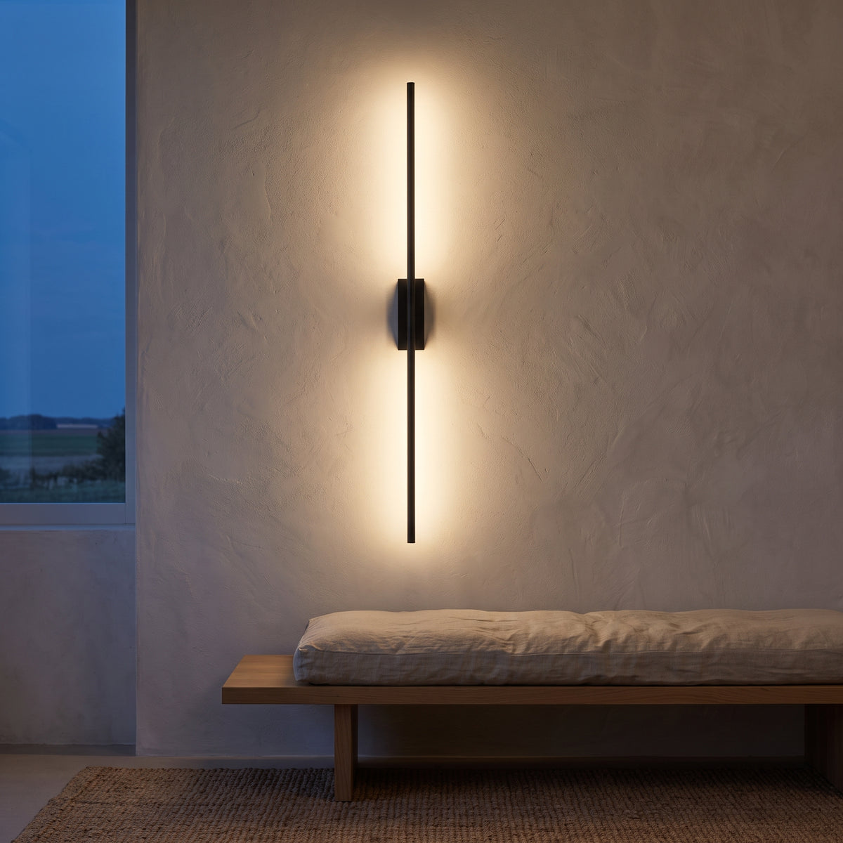Black minimalist LED bar wall sconce glowing on a plaster wall at dusk
