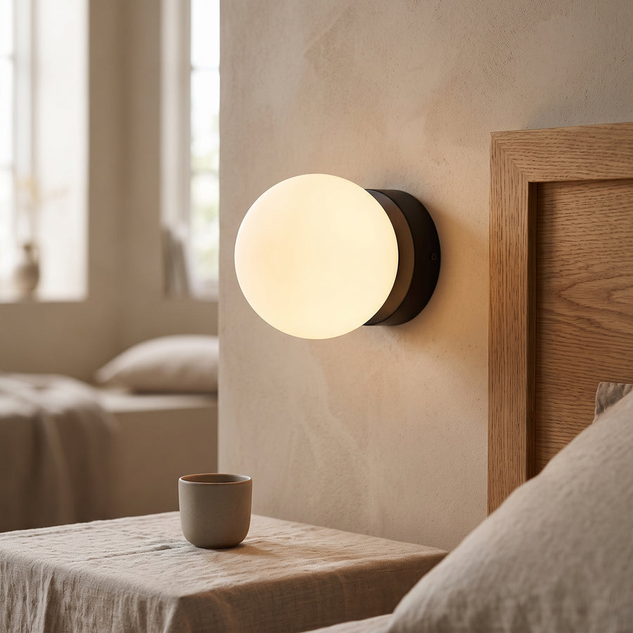 Opal glass globe wall light on a black disc glowing beside a bed