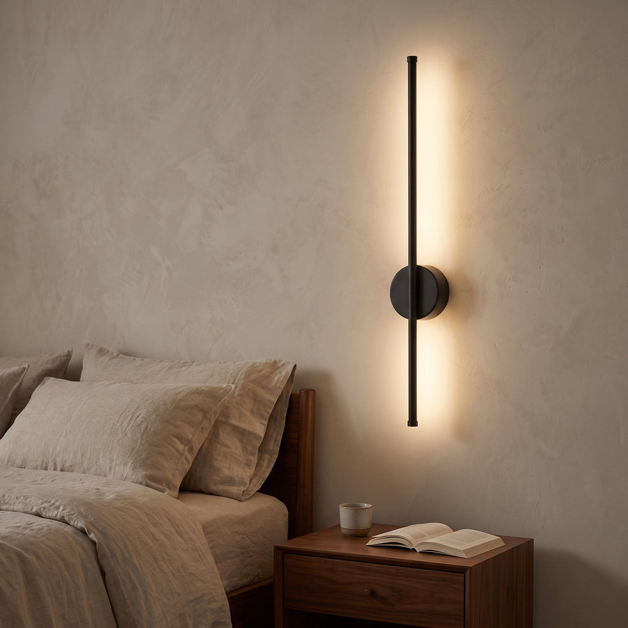Black round-plate rod wall sconce glowing beside a bedroom nightstand