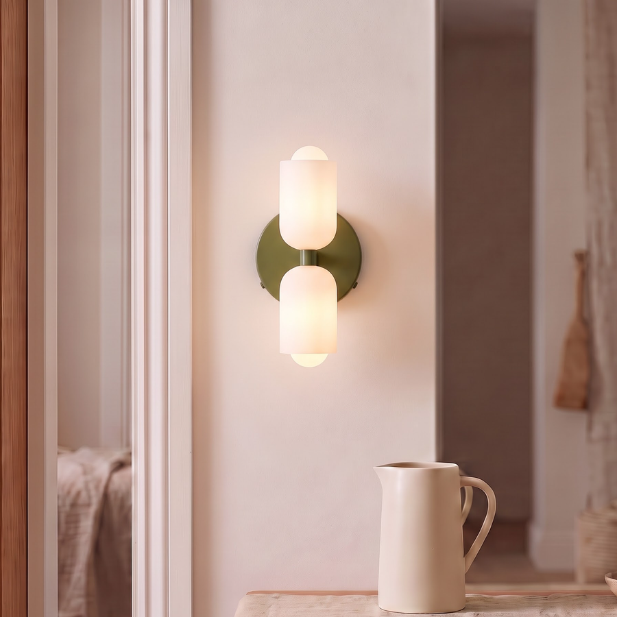 Olive green double capsule wall sconce glowing above a walnut nightstand