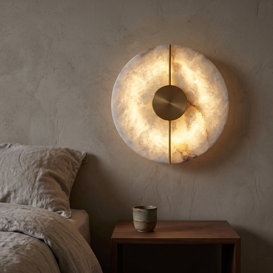 Round stone wall sconce glowing beside a linen bed at dusk